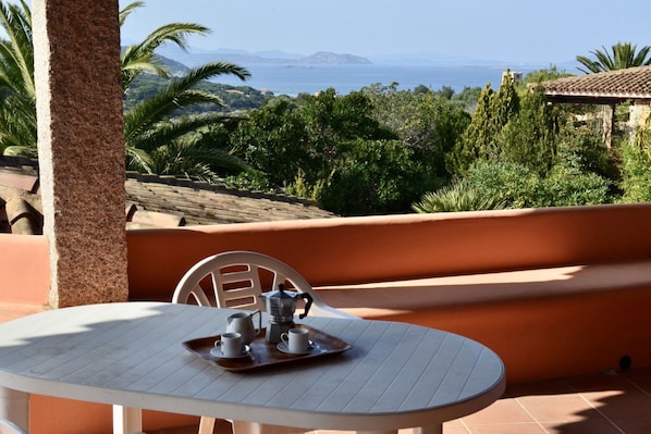 A terrace with a table set for two, overlooking a scenic landscape with mountains and the sea.