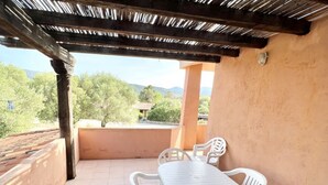 Un patio techado al aire libre con una mesa y sillas, vistas a árboles y montañas, y un edificio al fondo.