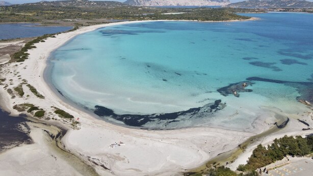 Vista aérea de una zona costera con una playa extensa de arena, aguas turquesas cristalinas y una montaña destacada al fondo.