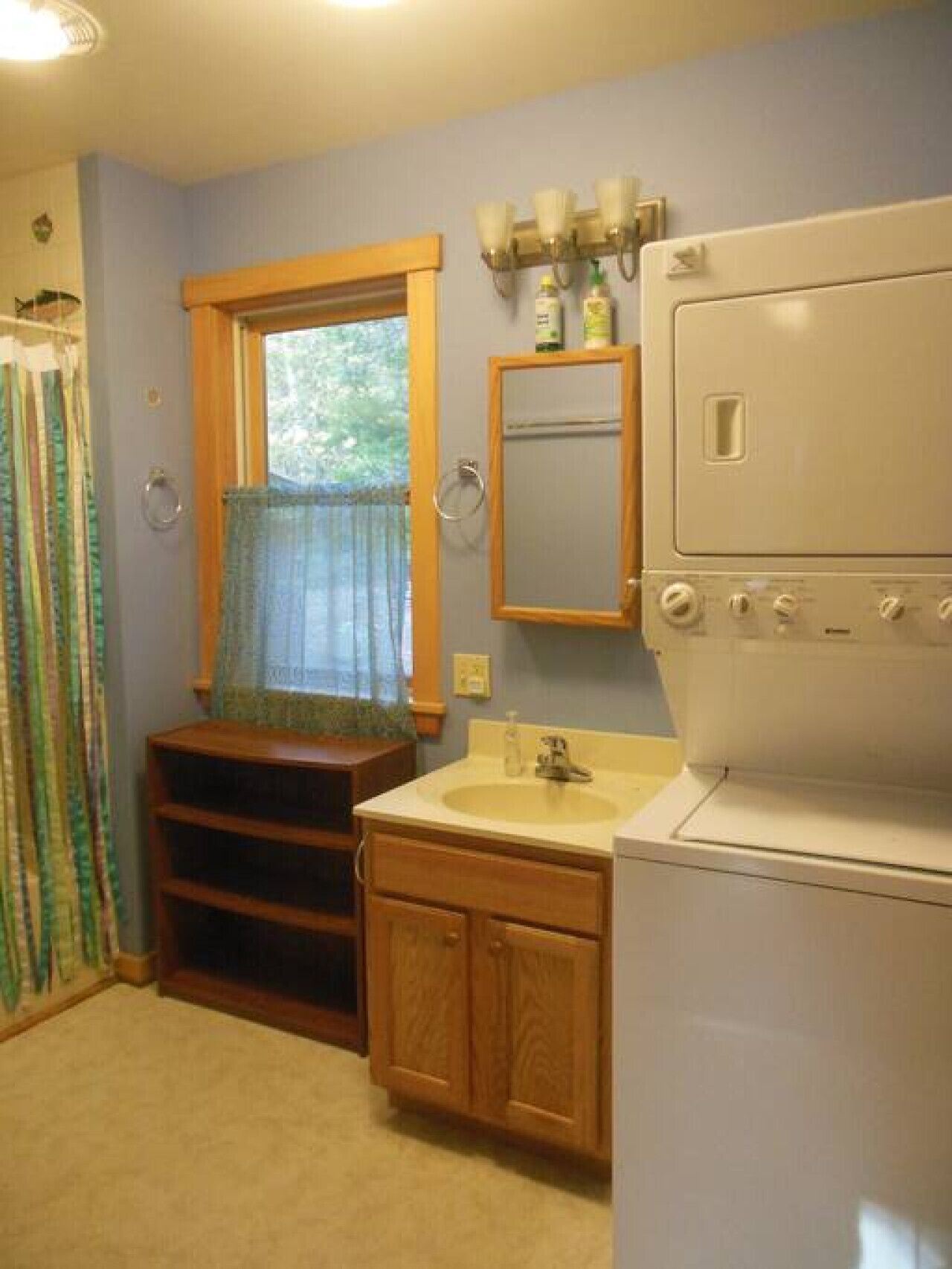 Un baño con lavamanos, espejo, ventana con cortinas y un electrodoméstico blanco.