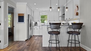 A modern kitchen with white cabinetry, a central island with bar stools, pendant lighting, and a view of the outdoors. - Happy Place! Golf Cart Included! (Emerald Isle)