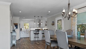 A spacious kitchen with white cabinets, a central island with stools, and a dining area with a wooden table and grey upholstered chairs. - Happy Place! Golf Cart Included! (Emerald Isle)