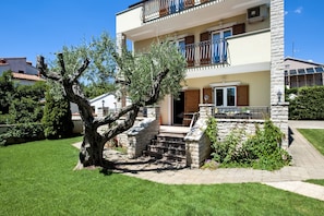 A two-story building with a stone staircase leading to the entrance, surrounded by greenery and a clear blue sky.