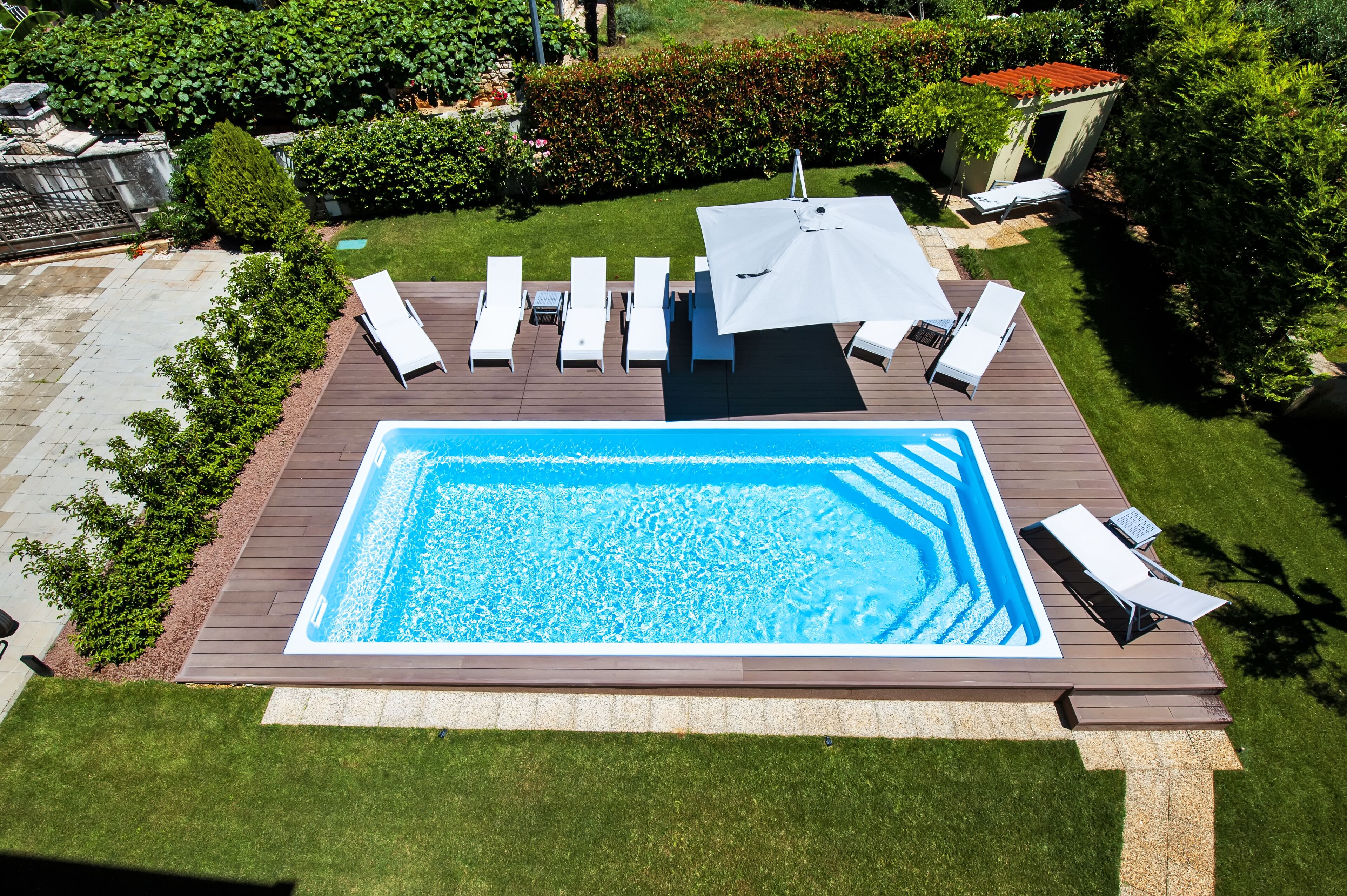 A rectangular swimming pool with a wooden deck, surrounded by white lounge chairs and a large umbrella.