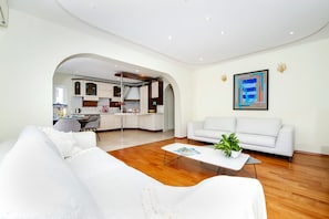 A modern living room with a white sofa, a coffee table, and a dining area with a kitchen visible through an archway.