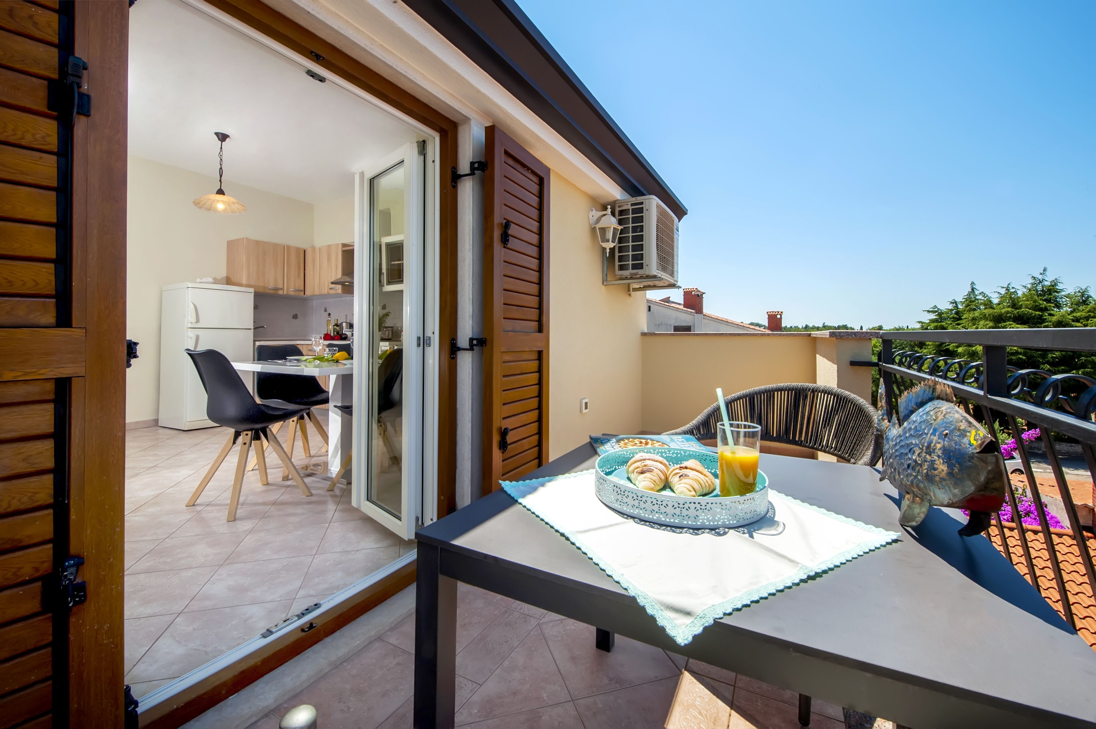 Un balcón con una mesa dispuesta para el desayuno, una cocina visible a través de una puerta abierta, y una escultura decorativa con forma de pez.