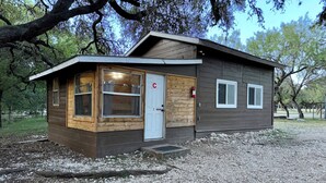 A small cabin with a wooden exterior, a white door, and a window with curtains. - Zubers - CA -C Cabin C (Concan)
