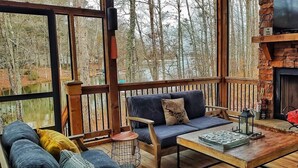 A cozy outdoor seating area with a stone fireplace, wooden deck, and a view of the forest. - Breathtaking porch just steps from the water. (Eatontonton)