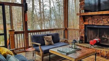 A cozy outdoor seating area with a stone fireplace, wooden deck, and a view of the forest.