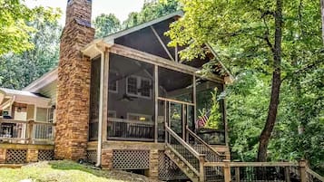 A cabin with a stone chimney, a covered porch, and a view of the surrounding forest.