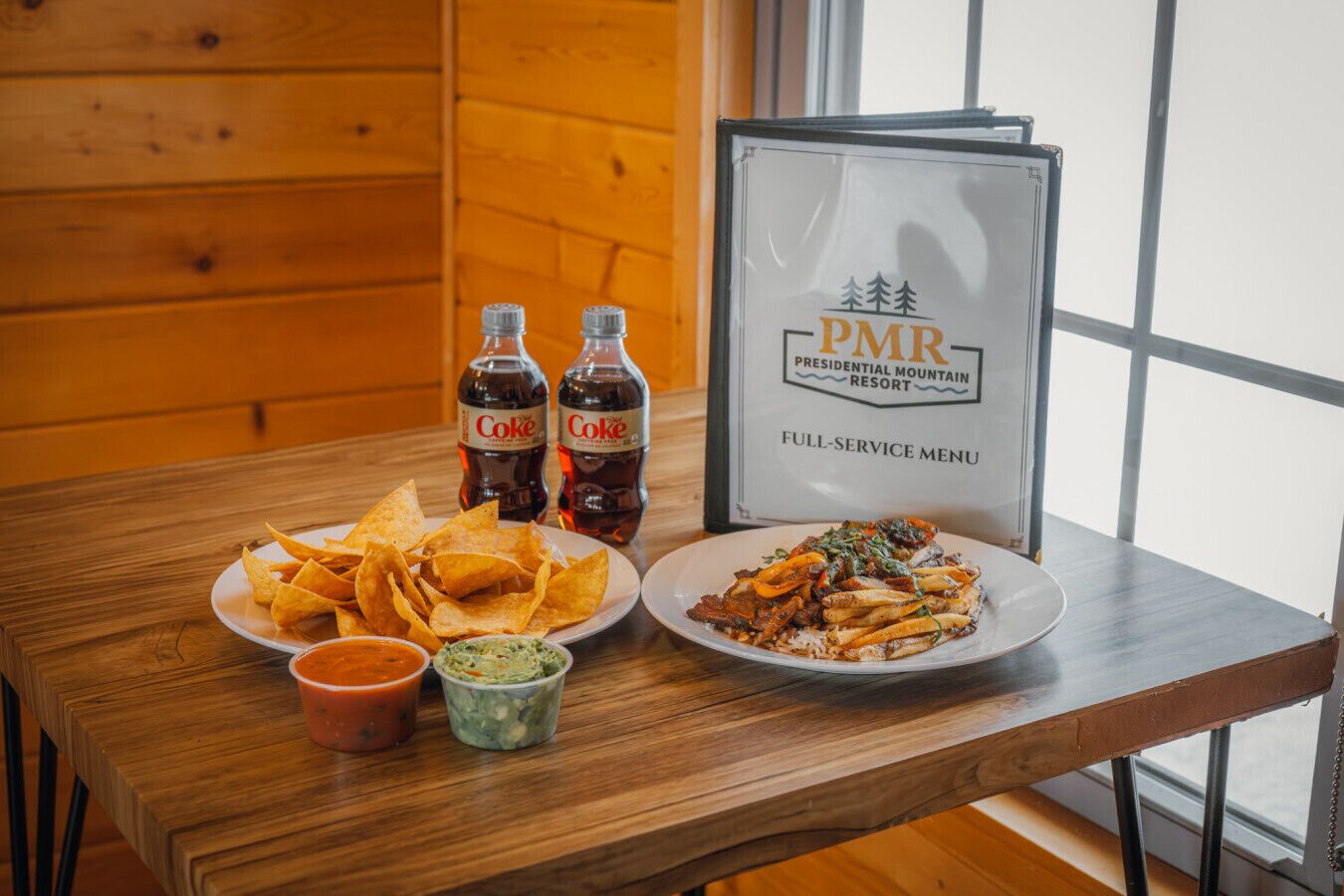 Ein Holztisch mit einer Nachos-Platte, zwei Cola-Flaschen, zwei kleinen Dip-Schalen und einer Speisekarte für das PMR Presidential Mountain Resort.