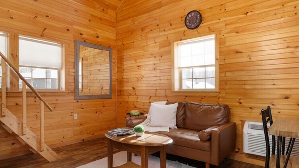 A cozy cabin interior with a wooden interior, a brown leather sofa, a small table with a plant, and a staircase. - Waterfront Log Cabin Hideaway Sauna Trails (Bethlehem)