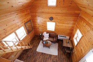 A cozy cabin interior with a wooden floor, a sofa, a small table, a window, and a staircase.