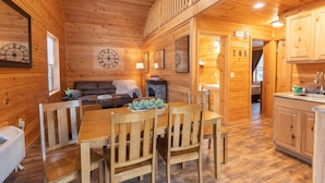 Interior de una cabaña de madera con mesa de comedor, sillas, sofá y zona de cocina.