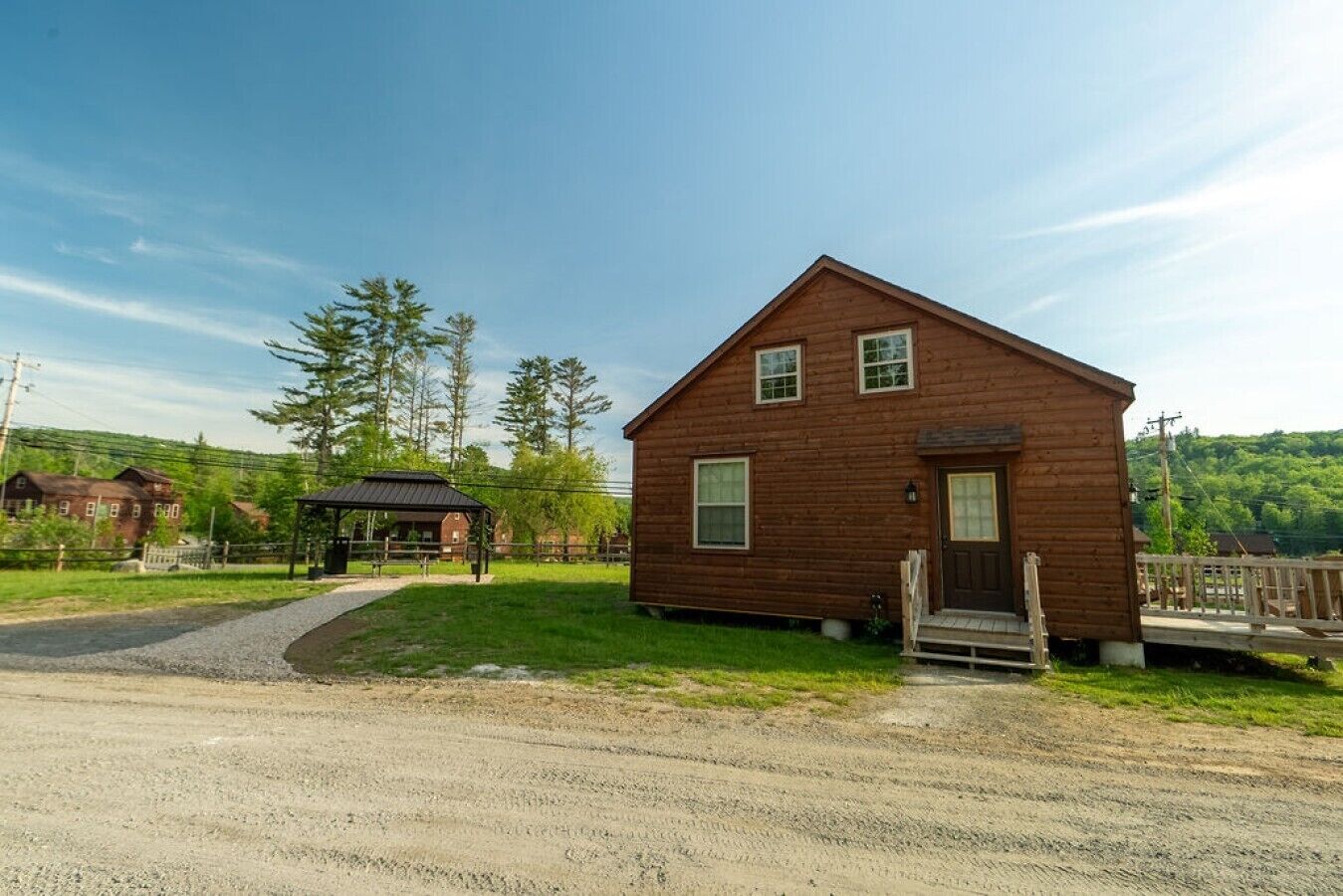 A small wooden cabin with a porch, a covered shelter, and a dirt road leading to it.