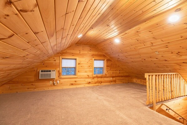 An attic room with wooden walls, two windows, an air conditioning unit, and a wooden staircase. - Two-Bedroom Log Cabin with Private Fire Pit (Bethlehem)