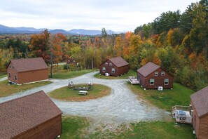 Un ensemble de cabanes en bois reliées par un chemin de terre, entourées d’un feuillage automnal et surplombées par des montagnes lointaines.