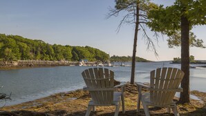 Zwei Adirondack-Stühle am Seeufer mit einem Steg und Booten im Hintergrund.