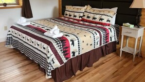 Quarto com piso de madeira, cama com colcha e almofadas estampadas, mesa de cabeceira com abajur e janela com cortinas.
