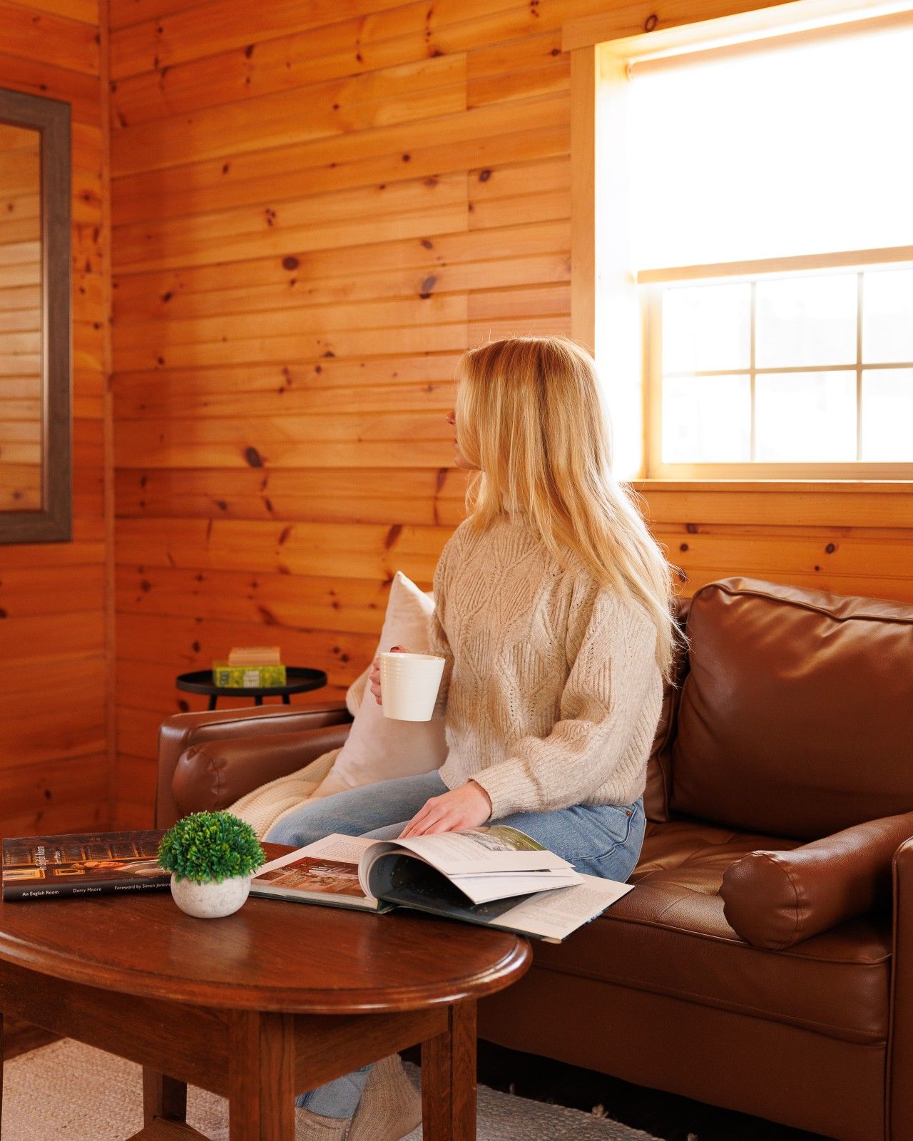A person sitting on a brown leather couch in a room with wooden walls, holding a white mug and reading a magazine.