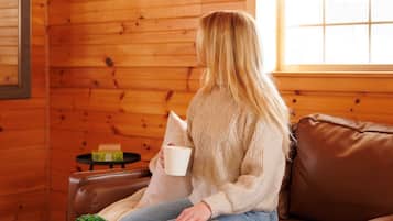 A person sitting on a brown leather couch in a room with wooden walls, holding a white mug and reading a magazine.