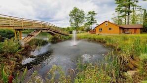 A wooden bridge over a pond with a fountain, surrounded by greenery and a wooden cabin.