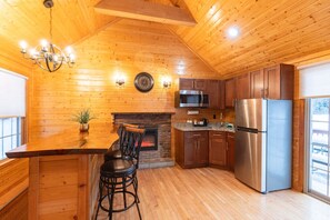 A cozy kitchen with wooden walls, a stone fireplace, stainless steel appliances, and a bar area with stools. - Serene Lakefront Cabin (Bethlehem)