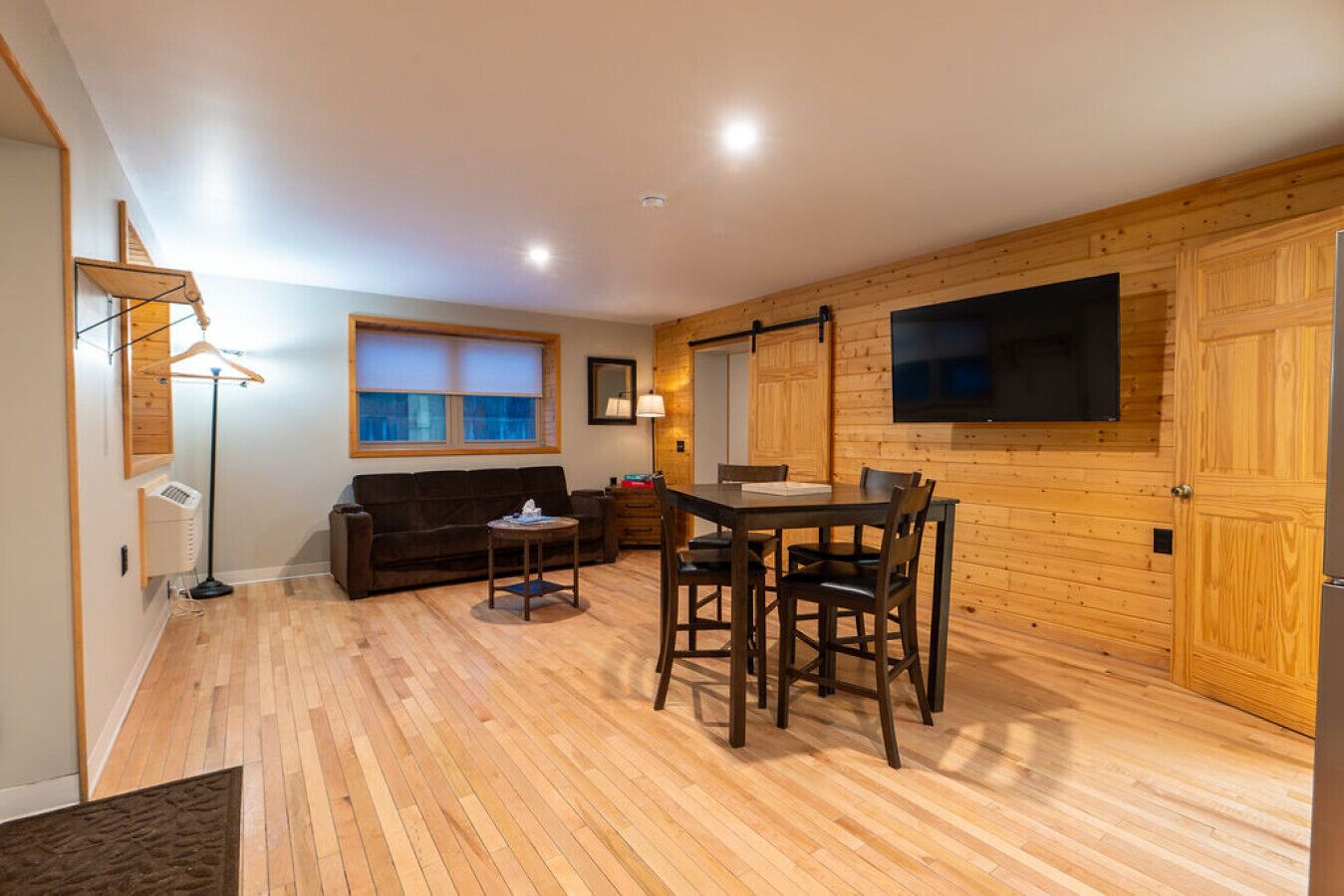 A cozy room with a wooden floor, a sofa, a dining table with chairs, a TV mounted on the wall, and a window with blinds.
