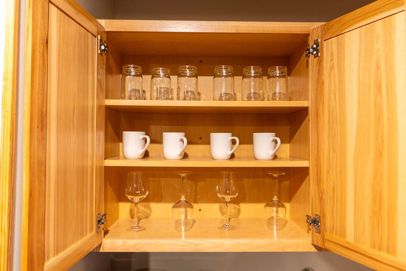A wooden cabinet with three shelves; the top shelf holds six glass jars, the middle shelf has four white cups, and the bottom shelf contains four wine glasses.