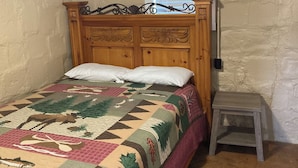 A bedroom with a wooden bed, a quilt, a small stool, and a window with blinds.