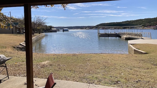 A covered outdoor area with a view of a lake, a dock, and a grassy area with a grill.