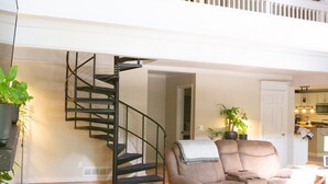 A spacious loft with a wooden floor, a beige sofa, a staircase, and a kitchen in the background. - A-Frame Arbor One Block From The Beach-Four Beds and Four Bathrooms! (Madison)