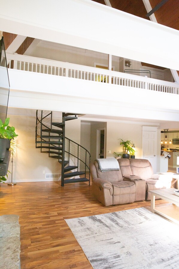 A spacious loft with a wooden floor, a beige sofa, a staircase, and a kitchen in the background.