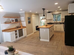 A modern kitchen with white cabinets, wooden countertops, and a central island. There is a refrigerator, sink, and a hanging light fixture.