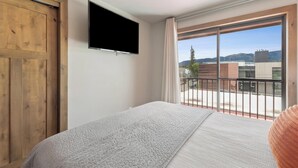 Ein Schlafzimmer mit Bett, einem an der Wand montierten Fernseher, einem Balkon mit Blick auf Gebäude und Berge sowie einer Holztür.