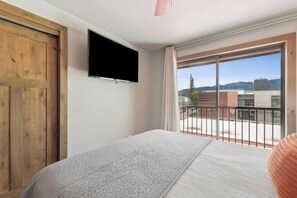 Ein Schlafzimmer mit Bett, einem an der Wand montierten Fernseher, einem Balkon mit Blick auf Gebäude und Berge sowie einer Holztür.