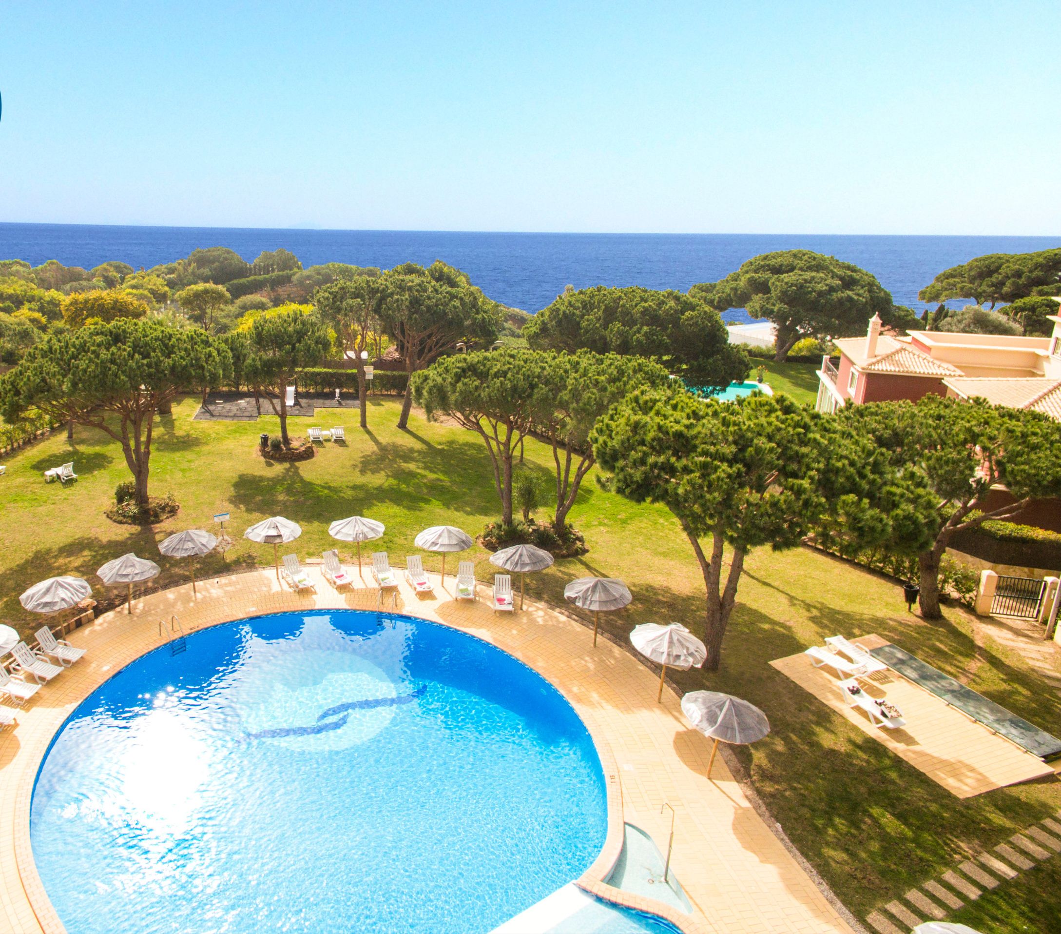A pool area with lounge chairs and umbrellas, surrounded by trees and a view of the sea.