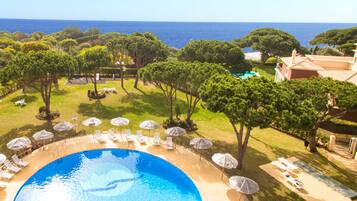 A pool area with lounge chairs and umbrellas, surrounded by trees and a view of the sea.