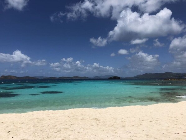 A beach with clear turquoise water, white sand, and distant mountains under a partly cloudy sky.