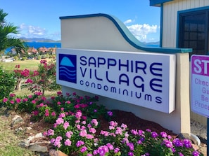 A sign for Sapphire Village Condominiums with a logo, surrounded by pink flowers and a clear view of the ocean.