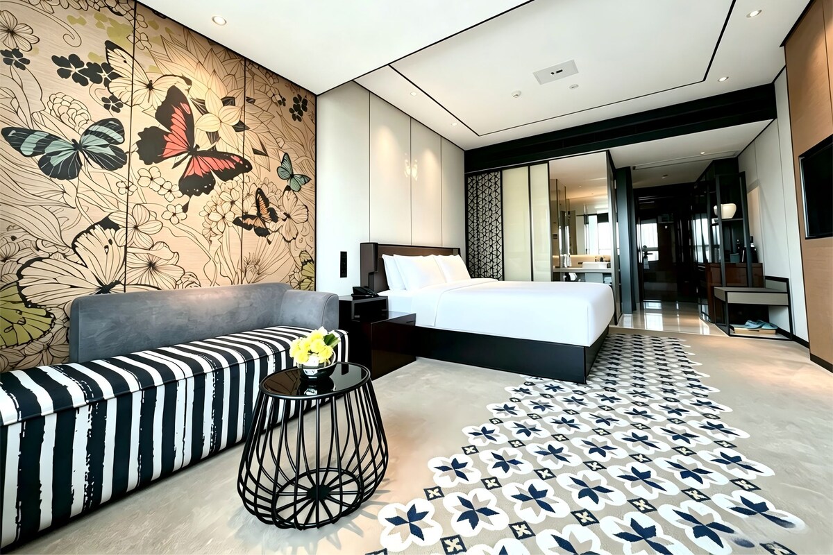 A modern hotel room with a large bed, a sofa, a small table with a vase, and a patterned floor.
