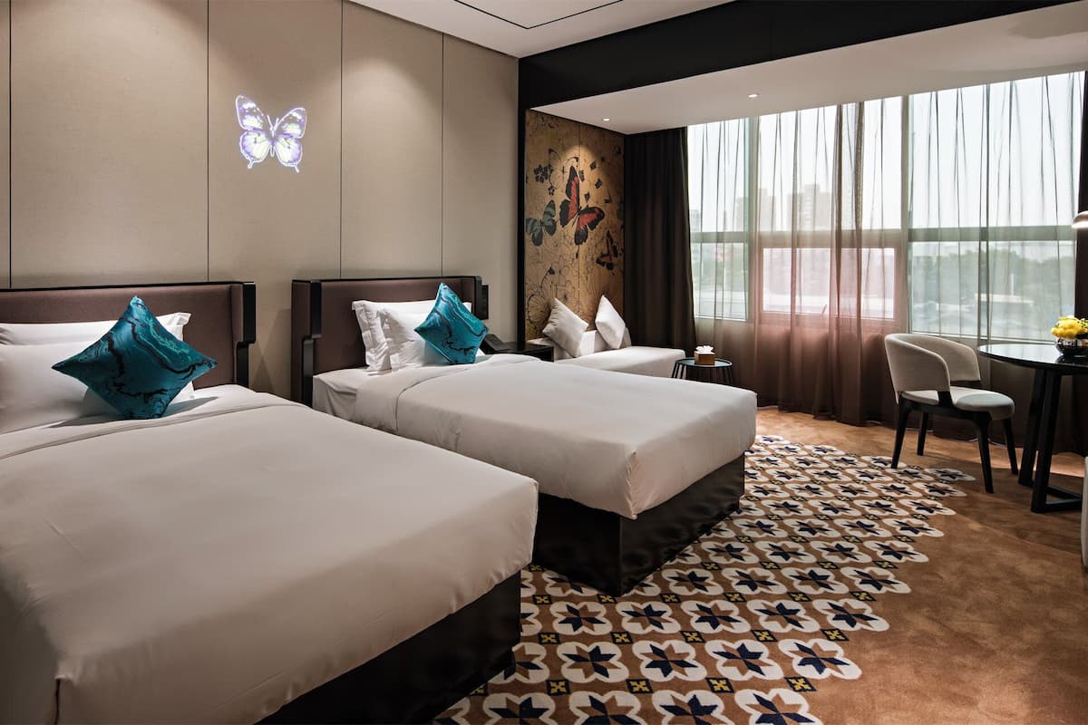 Habitación de hotel con dos camas, un escritorio, una silla y un ventanal con cortinas ligeras.