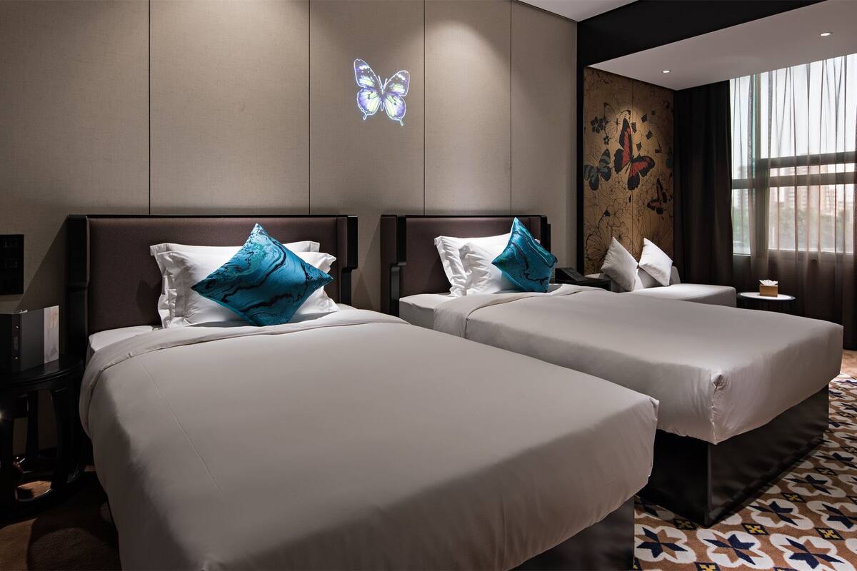 Habitación de hotel con dos camas, cada una con un colchón blanco y una almohada azul. Hay una lámpara de pared sobre las camas y un adorno de mariposa en la pared. La habitación tiene un ventanal con cortinas traslúcidas que dejan entrar luz natural.