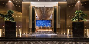 Un hall d’entrée moderne avec un comptoir d’accueil, un mur décoré de mosaïques bleues et deux arbres en pot.