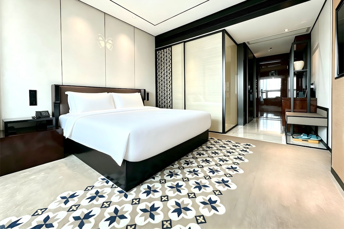 A modern hotel room with a large bed, a desk, and a view of the hallway.
