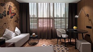 Ein Hotelzimmer mit Bett, einem Sessel, einem kleinen Tisch mit Lampe und Blick auf die Stadt durch große Fenster.