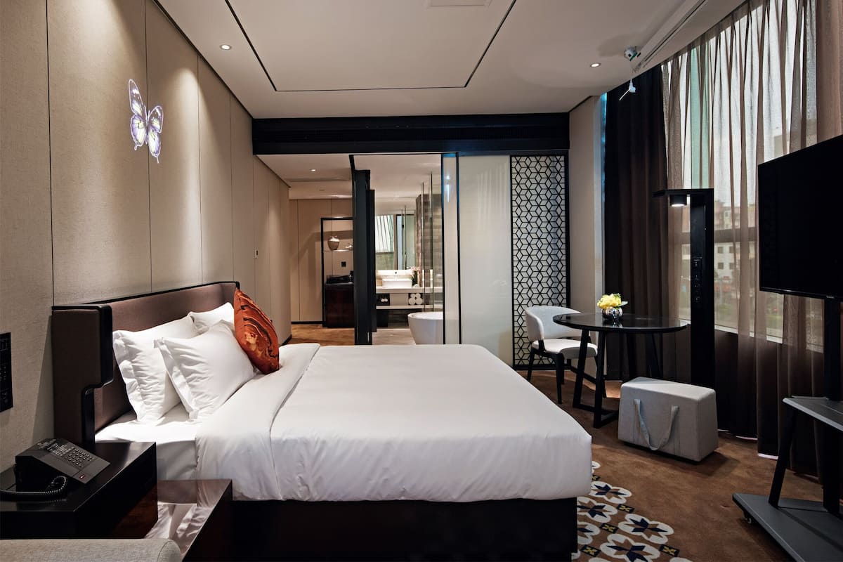 Una habitación de hotel moderna con una cama grande, mesita de noche y televisor. Hay un escritorio con una silla y una pequeña mesa con un florero. La habitación tiene un ventanal con cortinas y vistas a la ciudad.