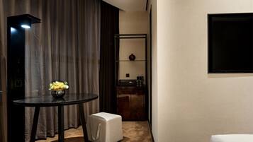 A hotel room with a round table, a small ottoman, a mirror, a flat-screen TV mounted on the wall, and a kitchenette area with a coffee machine.