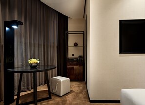 Une chambre d’hôtel équipée d’une table ronde, d’un petit pouf, d’un miroir, d’un téléviseur à écran plat fixé au mur et d’un coin cuisine avec une machine à café.