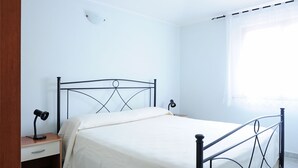 Een enkel bed met metalen frame, een nachtkastje met lamp en een raam met dunne gordijnen.